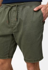 Shorts vert olive avec une taille élastique et un cordon de serrage. Comprend des poches latérales et une coupe décontractée avec des détails de coutures visibles.
