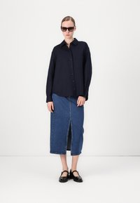 Marineblauwe knopsluiting overhemd met lange mouwen, gecombineerd met een spijkerrok met een voorklepsplit. Model draagt oversized zwarte zonnebril en zwarte schoenen.