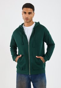 Hoodie zippé vert foncé, en coton, avec poches kangourou à l'avant, capuche avec cordon de serrage, porté sur un t-shirt blanc, associé à un jean bleu.