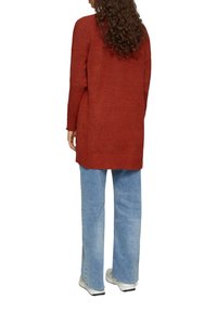 Pull long couleur rouille avec un tricot texturé, associé à un jean large bleu clair et des baskets blanches. Offre une coupe décontractée et un dos allongé.