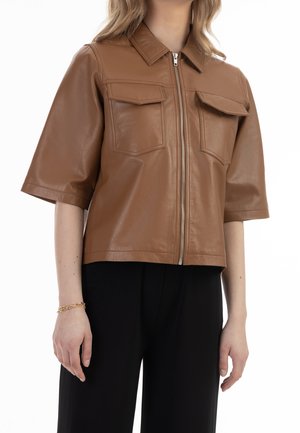 Femme portant une veste en cuir marron à manches courtes avec une fermeture éclair à l'avant et deux poches à rabat sur la poitrine, associée à un pantalon noir.