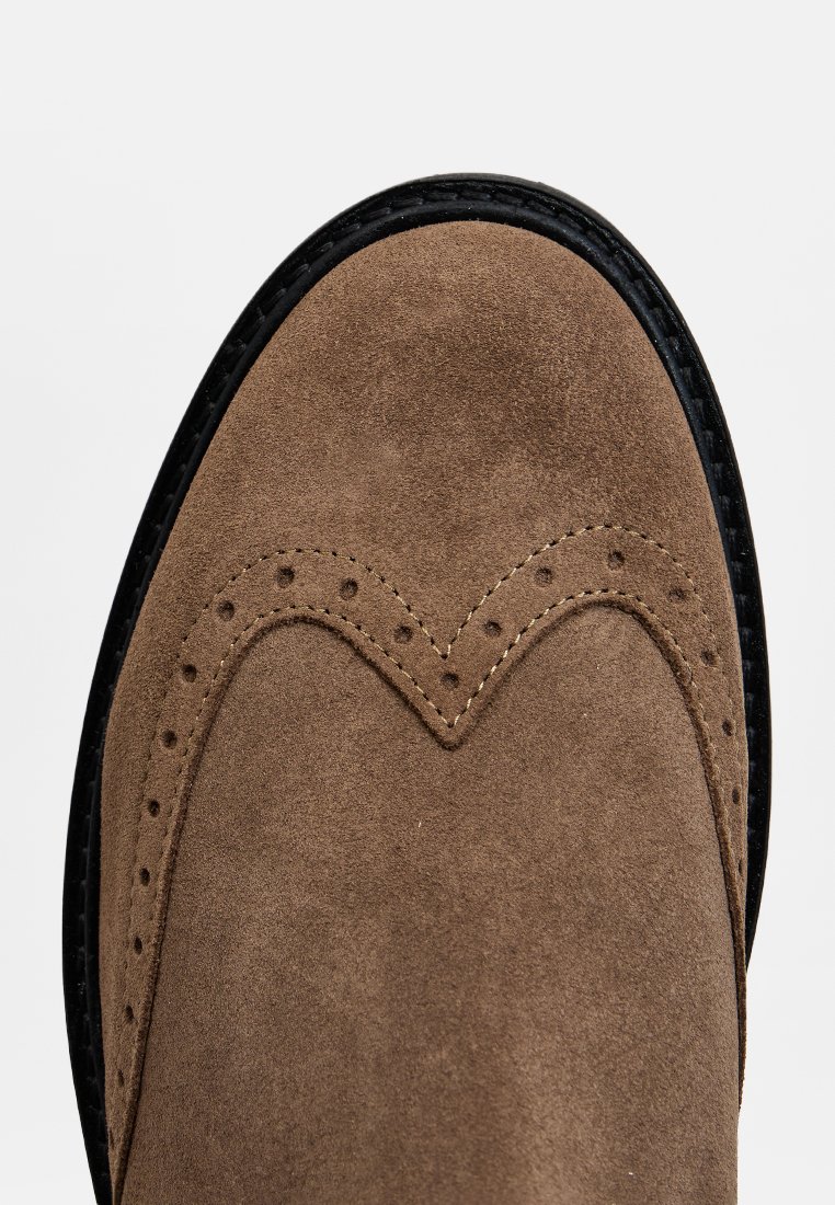 Braunes Wildleder-Schuh mit abgerundetem Zehenbereich, versehen mit dekorativen Perforierungen und einer kontrastierenden schwarzen Sohle. Glatte Textur mit genähten Akzenten.