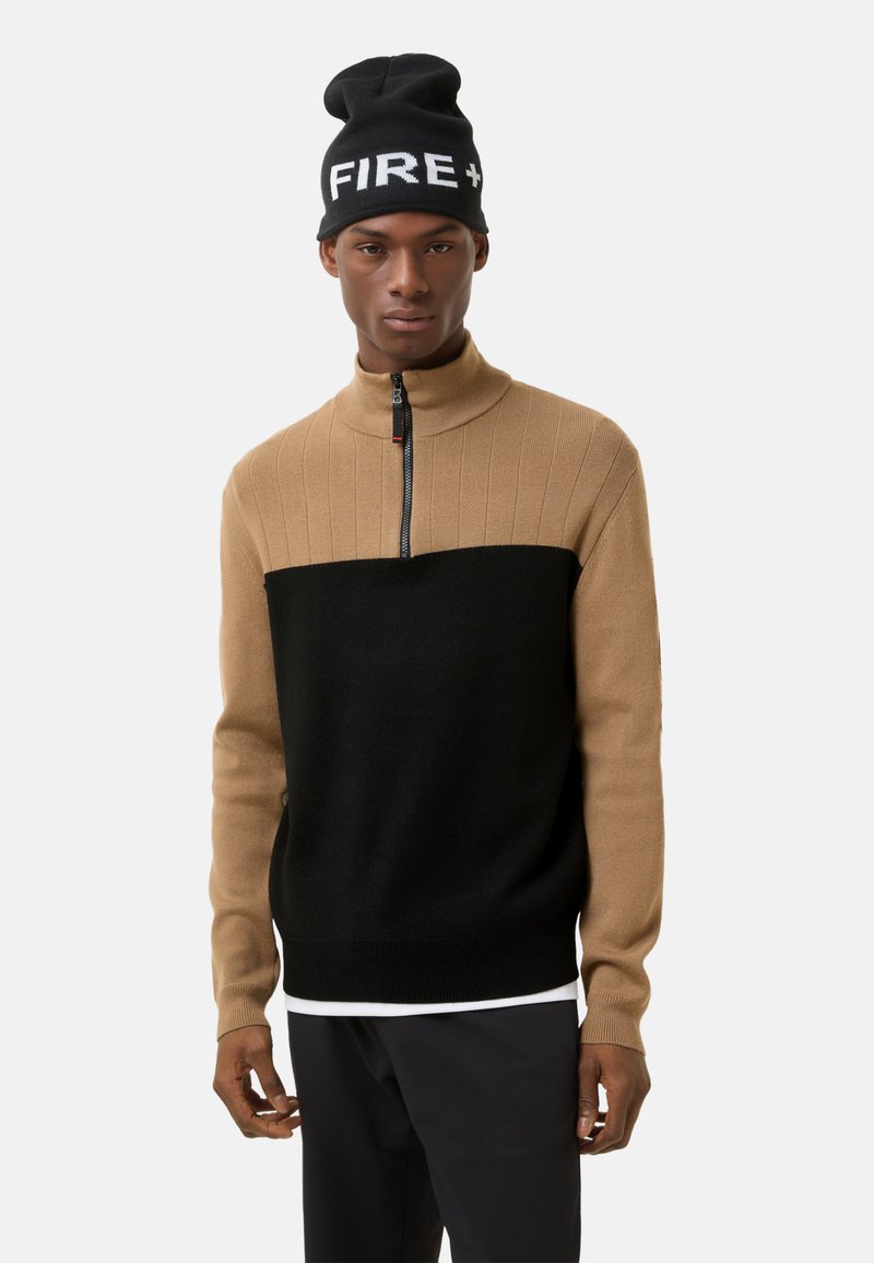 Pullover med høj krave, der har en lysbrun øverste del med ribbet struktur og en sort nederste del. Bæres med en sort beanie mærket "FIRE+".