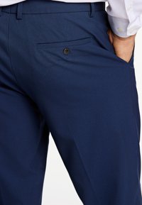 Spodnie w kolorze granatowym o gładkiej teksturze, z tylną kieszenią zapinaną na czarny guzik oraz kieszeniami bocznymi. Dopasowany krój dla efektu przylegającego.