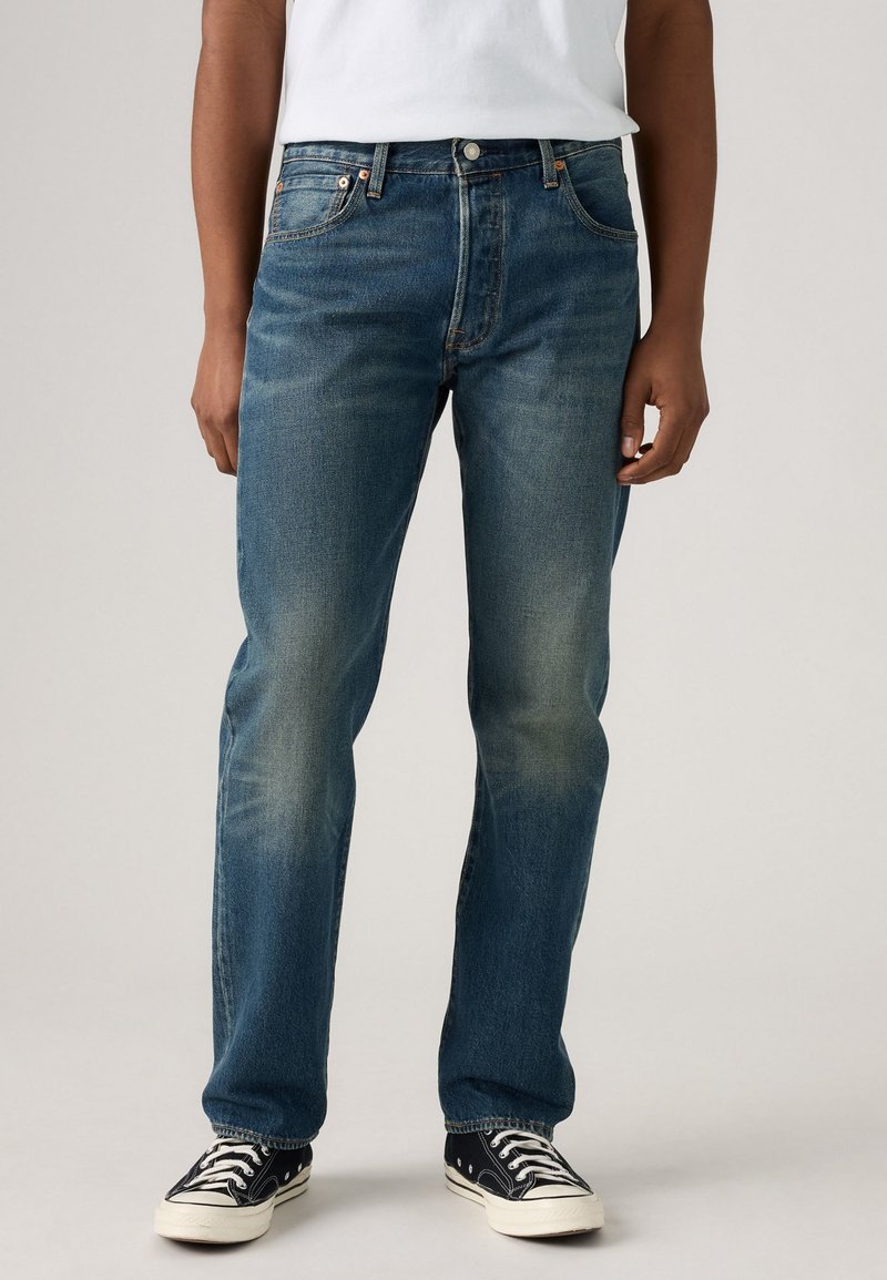 Blue denim jeans with a straight cut, faded texture, five pockets, and metal hardware. Paired with black and white sneakers.