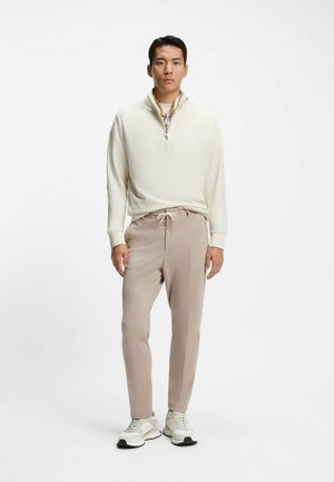 Hellbeige Zip-up-Pullover mit strukturierter Oberfläche, kombiniert mit sanft beigen, schmal zulaufenden Hosen und hellen Sneakern mit dunklen Außensohlen.