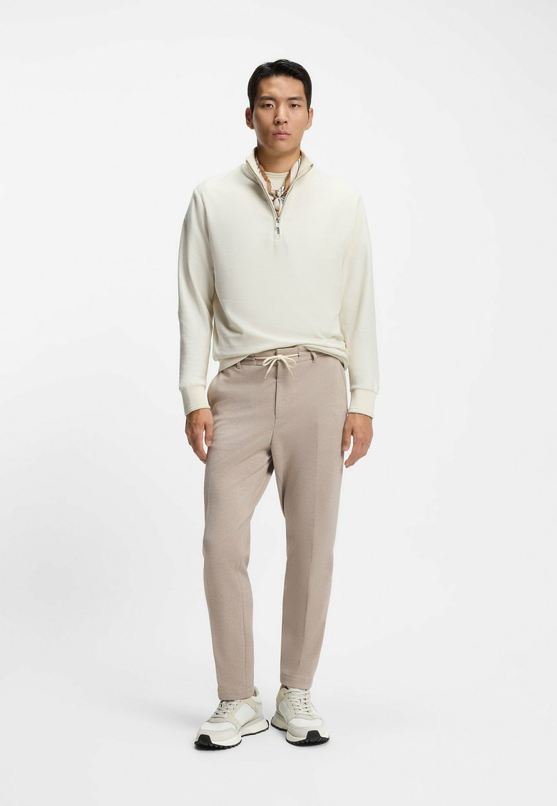 Maglione zip-up color crema chiaro con finitura testurizzata abbinato a pantaloni tapered beige morbidi e sneakers chiare con suole scure.