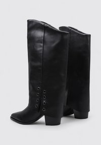 Botas de piel negra hasta la rodilla con punta afilada, presentando una textura suave y ojales metálicos decorativos en el lateral. Diseño de tacón cuadrado.