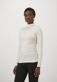 Langärmliges, hochgeschlossenes Oberteil aus hellbeigem Stoff mit taillierter Silhouette und gerafften Details an den Seiten, kombiniert mit schwarzen Hosen.