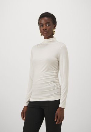 Long-sleeve, high-neck top in light beige fabric with a fitted silhouette and gathered detailing at the sides, paired with black pants.