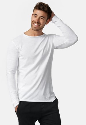 Jeune homme aux cheveux courts et foncés souriant, portant une chemise blanche à manches longues et un pantalon noir, une main touchant sa tête.