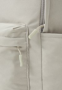 Sac à dos en tissu gris clair avec une surface texturée, équipé d'une fermeture éclair et de deux languettes en tissu attachées à des poches inclinées.
