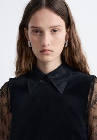 Schwarzes ärmelloses Top mit strukturiertem Kragen, texturiertem Stoff, über einem transparenten schwarzen Spitzenlangarmhemd getragen. Silberne Creolen.