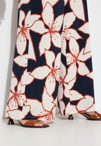 Námořnické široké kalhoty s velkým bílým květinovým vzorem doplněným červenou, v kombinaci s hnědými sandály na podpatku s řemínky a krokodýlí texturou.