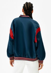 Granatowa satynowa kurtka bombowa z czerwonymi akcentami na ramionach. Posiada ściągacz na kołnierzu i mankietach w wielokolorowe paski oraz elastyczny pas.