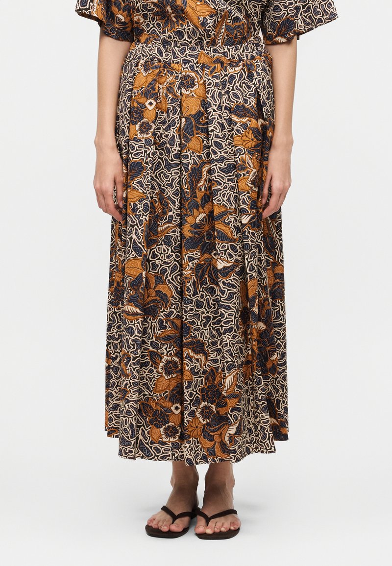 Γυναίκα που φορά μακριά φλοράλ φούστα με πορτοκαλί, μπλε σκούρο και μπεζ χρώματα και μαύρα σαγιονάρες, στέκεται μπροστά σε μονόχρωμο φόντο.