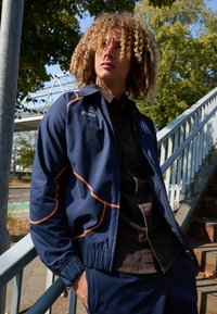 Mornarsko modra jakna z oranžnimi šivi, izdelana iz lahkega materiala. Ima visok ovratnik in elastične manšete, nosi se preko črne majice.