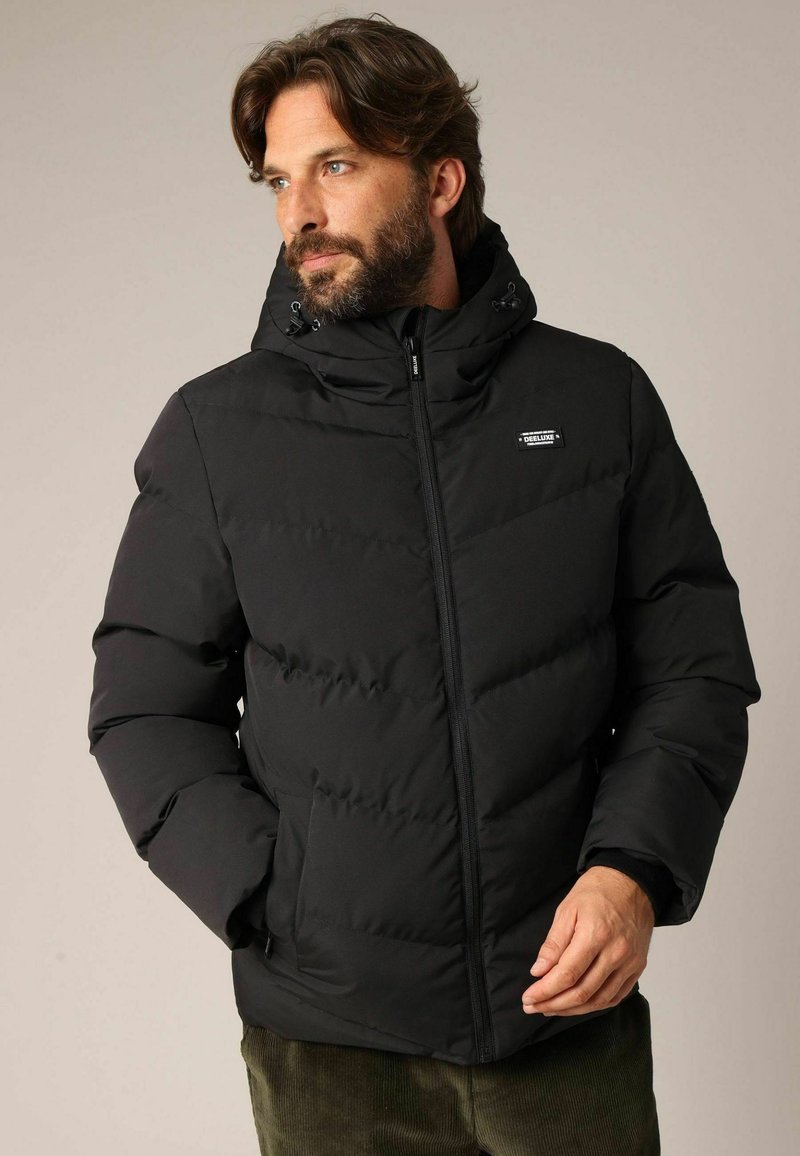 Veste matelassée noire avec fermeture éclair à l'avant, large capuche, coutures diagonales et un emblème sur le côté gauche de la poitrine. Matériau doux et isolant.