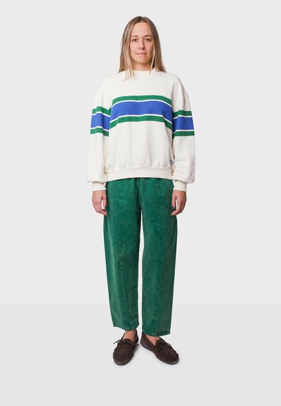 Crème sweatshirt met blauwe en groene strepen, gecombineerd met groene textuurbroek. Donkerbruine zachte schoenen maken de outfit compleet.