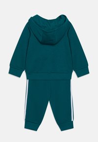 Tuta teal composta da una felpa con cappuccio e polsini a costine, abbinata a pantaloni con strisce bianche sui lati. Realizzata in un morbido tessuto elasticizzato.