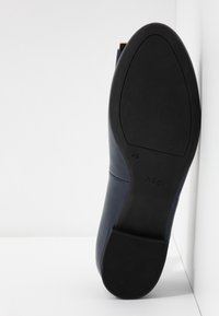 Semelle d'une chaussure habillée bleu marine inclinée contre un coin blanc, dotée d'un talon en caoutchouc et de la marque "hōgl" sur la semelle.