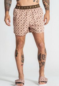 Costumi da bagno rosa con un motivo a logo marrone ripetuto, vita elastica e una vestibilità rilassata. Abbinati a sandali a scivolo rosa chiaro.
