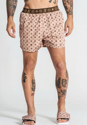 Bañador rosa con un patrón repetido de logo marrón, cinturilla elástica y un corte holgado. Combinado con sandalias de playa rosa claro.