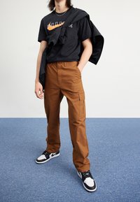 Bruna cargobyxor med sidofickor, tillsammans med en svart t-shirt som har en vit och orange logotyp, samt svarta och vita sneakers.