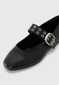 Chaussure en cuir noir de style Mary Jane avec un bout carré, comportant une sangle ornée de rivets en argent et une boucle en métal rectangulaire.