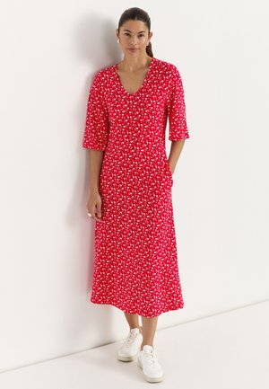 Femme aux cheveux noirs attachés en queue de cheval, portant une robe midi rouge à imprimé floral blanc et des baskets blanches, debout contre un mur blanc.