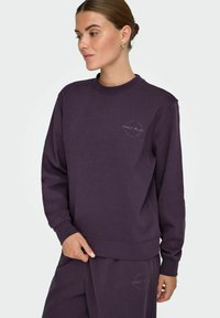 Lilla sweatshirt med rund halsudskæring, lange ærmer og ribbede manchetter. Har et cirkulært logo print på venstre bryst. Blød stofstruktur.