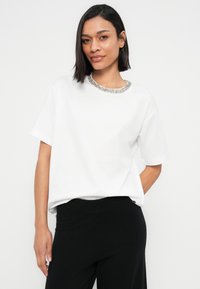 Λευκό πουκάμισο με κοντά μανίκια και λαιμόκοψη διακοσμημένη με χάντρες. Μαλακό ύφασμα, άνετη εφαρμογή και απλός σχεδιασμός. Φορεμένο με μαύρο παντελόνι.