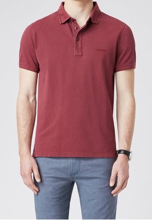 Polo color marrone in cotone, con pattina a tre bottoni e maniche corte. Indossata con jeans azzurri. Design semplice, vestibilità comoda.