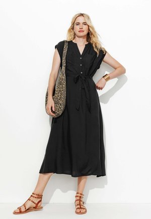 Femme blonde en robe midi noire boutonnée avec ceinture à nouer, tenant un sac à épaule imprimé léopard, portant des sandales marron cloutées et un bracelet manchette doré.