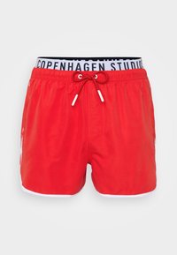 Red swim shorts with white trim, elastic waistband featuring "COPENHAGEN STUDIOS" text, adjustable drawstring with white tips.