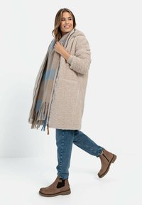 Beiger, flauschiger Mantel mit Kapuze, leichter Schal mit blauem und beige kariertem Muster, blaue Jeans, braune Stiefeletten, auffällige Taschen.