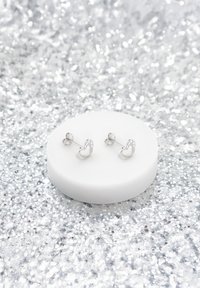 Silver hjärtformade stickörringar med en slät yta och små klara detaljer, uppställda på en vit cirkulär ställning mot en glittrig bakgrund.