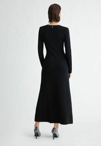 Schwarzes langärmliges Kleid mit tailliertem Schnitt und ausgestelltem Rock. Verfügt über einen runden Rückenausschnitt mit einem kleinen Logodetail. Gemusterte High-Heels.