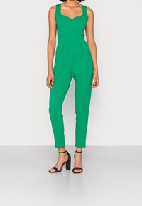Green sleeveless jumpsuit with a sweetheart neckline, fitted bodice, and tapered legs. Worn with black block-heeled sandals.