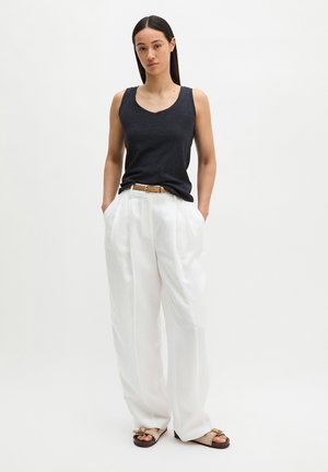 Frau mit langen dunklen Haaren trägt ein schwarzes ärmelloses Oberteil, weiße, hoch taillierte, weit geschnittene Hosen, einen hellbraunen Gürtel und beige Sandalen und steht mit den Händen in den Taschen.