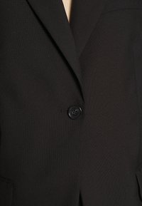 Närmsta bild av en skräddarsydd svart kavaj som är knäppt i mitten, vilket visar den texturerade tyget och en del av slaget och kragen.