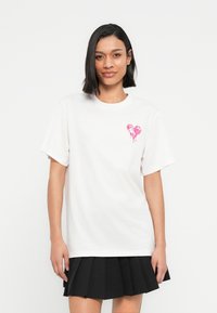 Λευκό βαμβακερό t-shirt με ροζ καρδιά στο αριστερό στήθος. Κοντά μανίκια και στρογγυλή λαιμόκοψη. Απλή, άνετη γραμμή.