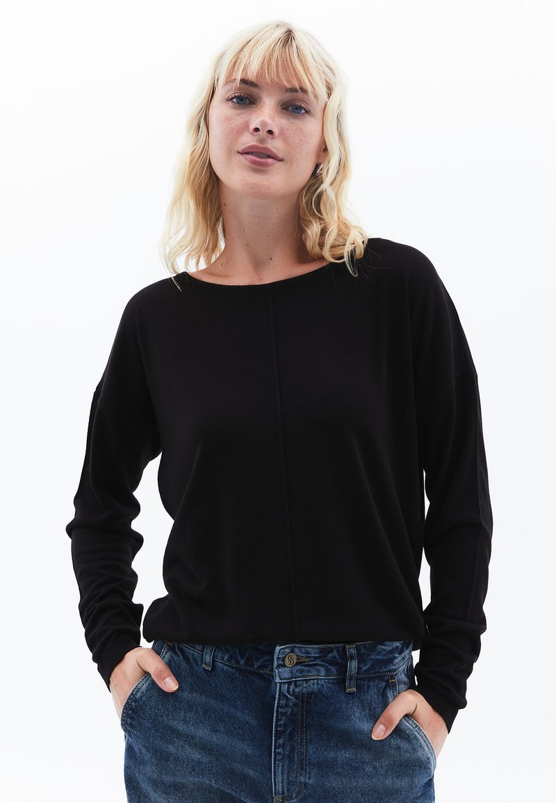 Top nero a maniche lunghe con scollatura a barchetta e spalle cadenti, abbinato a jeans in denim blu a vita alta con dettaglio della chiusura a bottone.