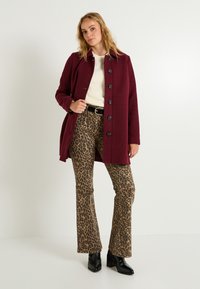 Abrigo de lana burdeos con botones negros, combinado con pantalones acampanados de estampado de leopardo marrón y botas de tobillo negras con tacón. Tela texturizada visible.