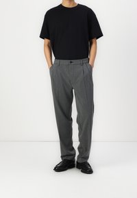 T-shirt nera con vestibilità rilassata, pantaloni grigi a righe con chiusura a bottoni, fronte plissettato, tasche laterali e scarpe nere.
