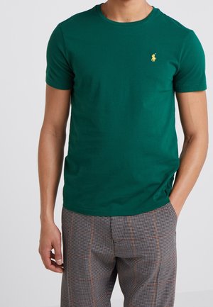 Man wearing a plain dark green short-sleeve t-shirt with a small yellow logo on the chest and checked trousers, standing with one hand in pocket.
