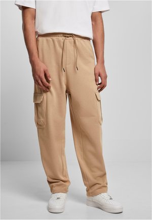 Homme portant un pantalon de jogging cargo beige avec taille à cordon et des baskets blanches, debout devant un fond gris uni.