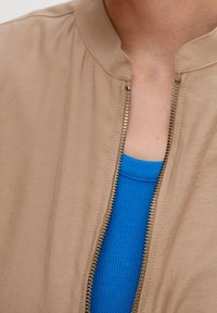 Tan bomberjakke med høj krave, guldlynlås og en ribbet blå tanktop under. Glat tekstur med et minimalistisk design.