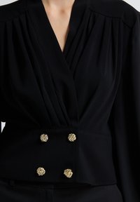 Sort bluse med plisserede skuldre, omslag design og gyldne blomsterknapper i taljen. Blødt stof med en glat tekstur.