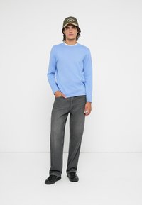 Suéter de mangas compridas azul-claro com decote branco, combinado com jeans cinza-escuro de perna reta e sapatos pretos; modelo usa um boné oliva.
